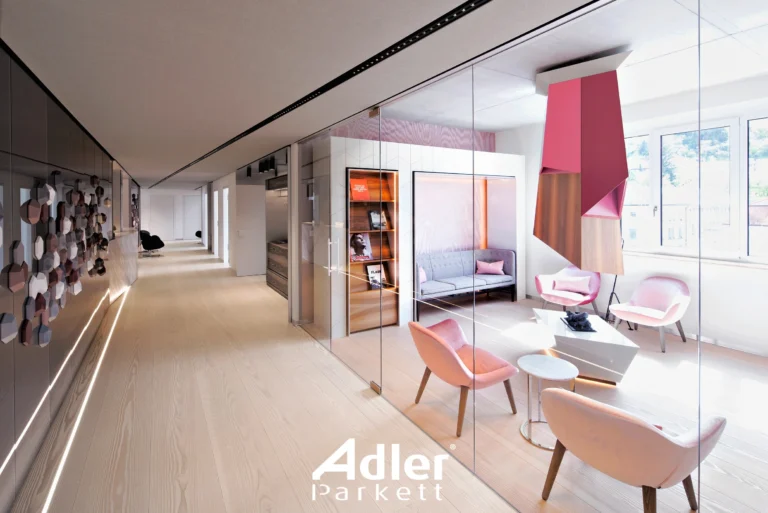 Moderner Bürobereich mit hellem Adler Parkett, Glaswänden und pinken Akzenten. Offener Flur und Sitzgelegenheiten.