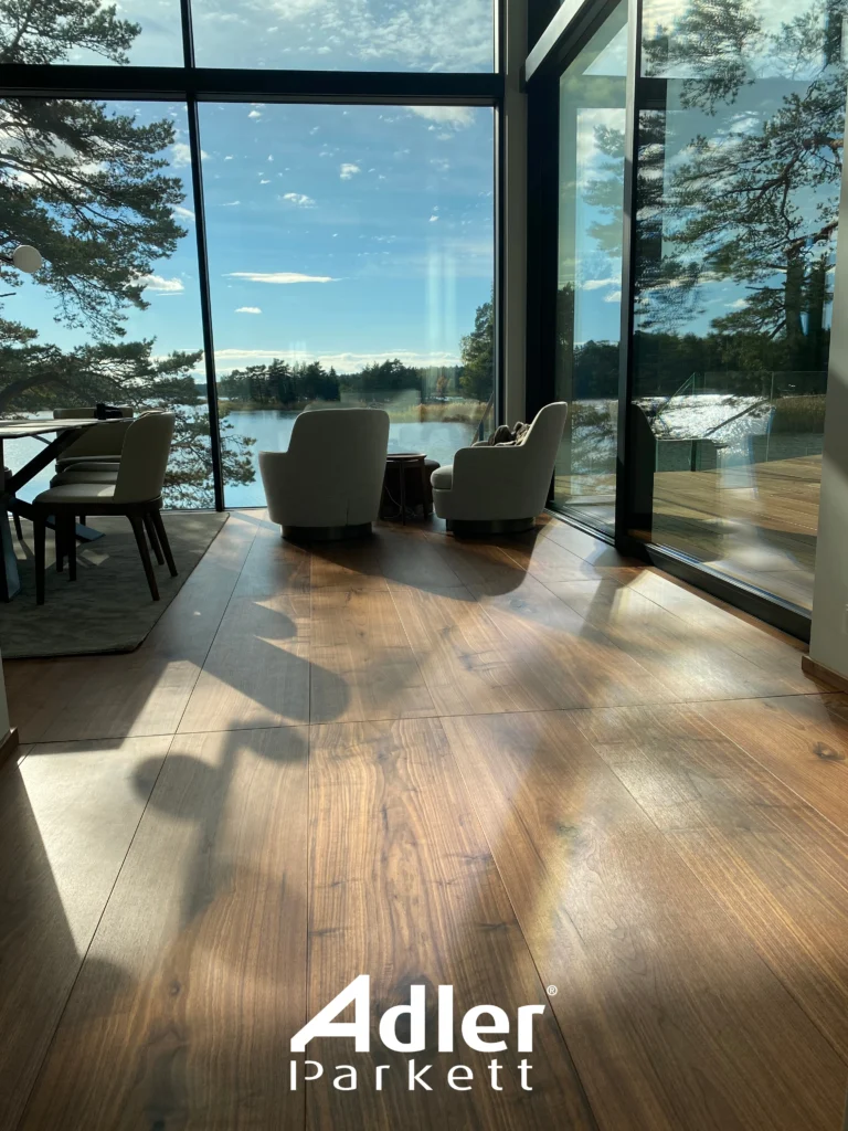 Raum mit Adler Parkett, bodentiefen Fenstern und Blick auf einen See. Helle Sessel laden zum Entspannen ein.