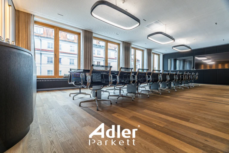 Großer Konferenzraum mit Adler Parkett, langer Tischreihe, vielen Stühlen und großen Fenstern. Moderne Deckenleuchten.