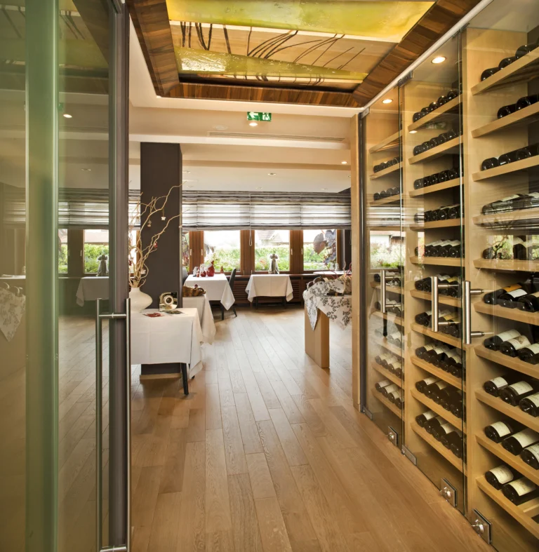 Restaurant mit hellbraunem Holzboden und einglasertem Weinkeller an der Seite. Tische mit weißen Tischtüchern und Fenstern.