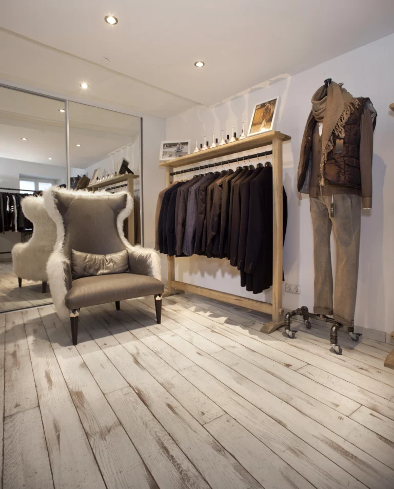 Innenansicht eines Bekleidungsgeschäfts mit weiß gekalktem Holzboden, Kleiderstange, Schaufensterpuppe und einem großen pelzigen Sessel.