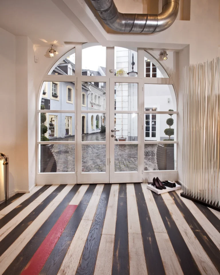 Eingangsbereich mit auffälligem schwarz-weiß gestreiften Holzboden mit rotem Akzent. Großes Rundbogenfenster mit Blick auf einen Hof.