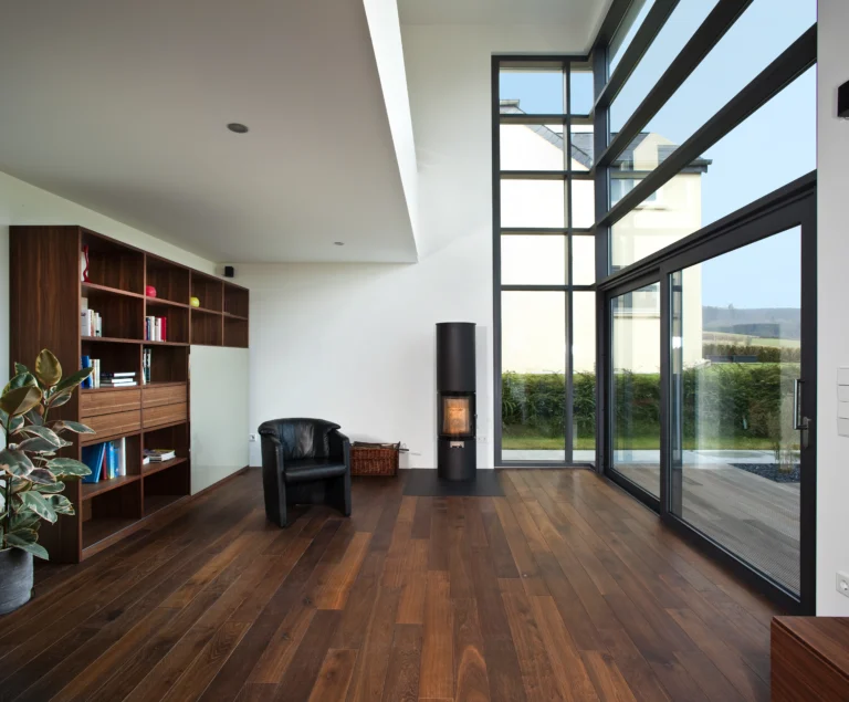 Helles Wohnzimmer mit dunklem "Vieux de France" Holzboden, einer Bücherwand, schwarzem Sessel und hohen Fenstern mit Blick ins Grüne.