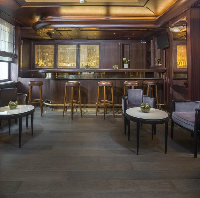 Eine klassische Hotelbar mit dunklem Holz. Der Boden ist ein grauer Holzdielenboden. Barhocker, Tische und Sessel runden das Bild ab.