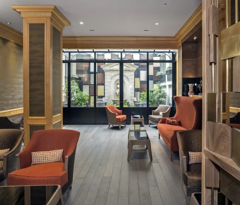 Eine elegante Hotel-Lounge mit hellem Holzdielenboden. Rote Sessel und Sofas. Große Glasfront mit Blick auf einen Innenhof mit Brunnen.